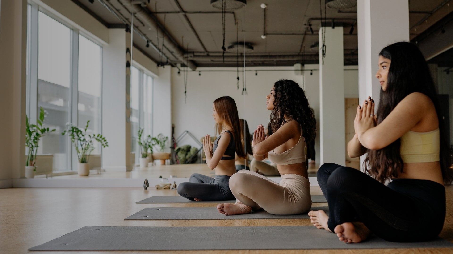 Three women practicing yoga in a studio with large windows and plants.